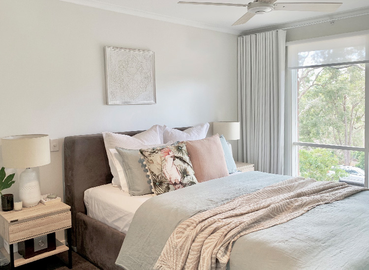 A beautiful bedroom with white blockout curtains and a sunscreen roller blind looking out to a garden and gum trees.