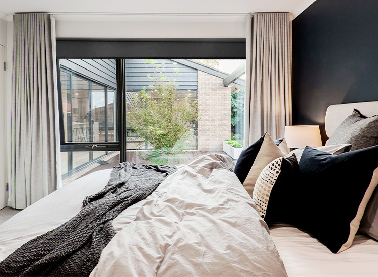 A dramatic and modern bedroom featuring a large open window to a courtyard, with grey blockout curtains over a black blockout blind.
