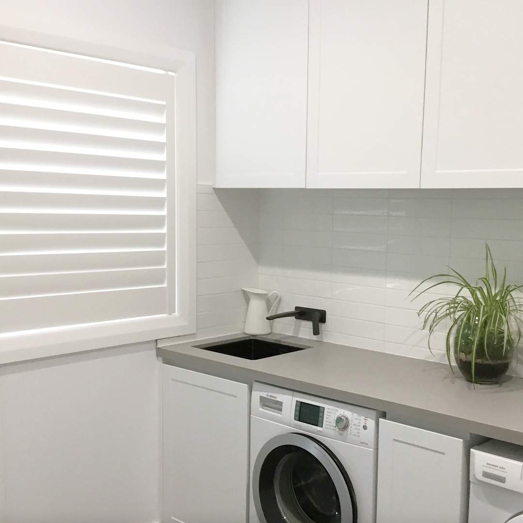 Sleek white laundry with overhead cupboards and a white plantation shutter on the window