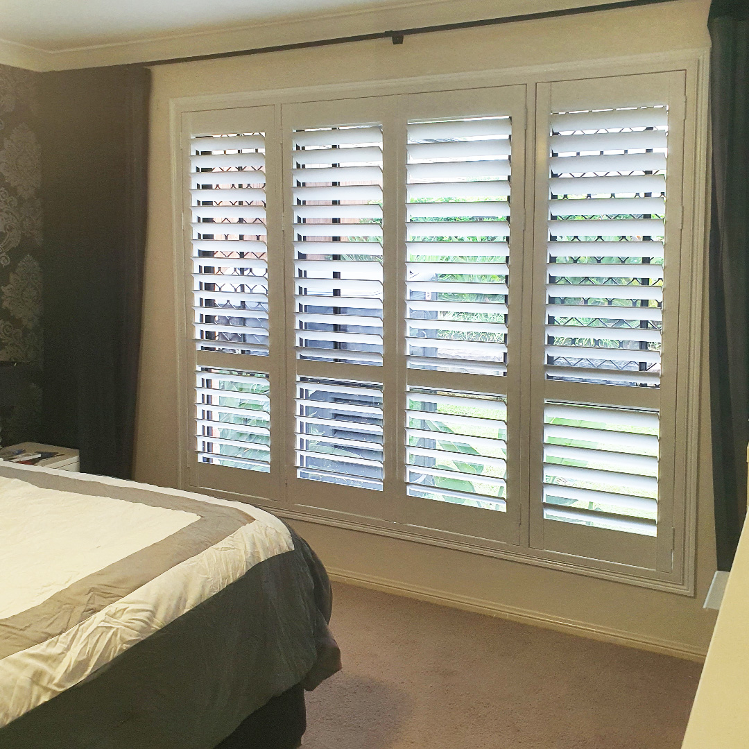 Large window in a main bedroom with a white plantation shutter and dark sheer curtains on it.