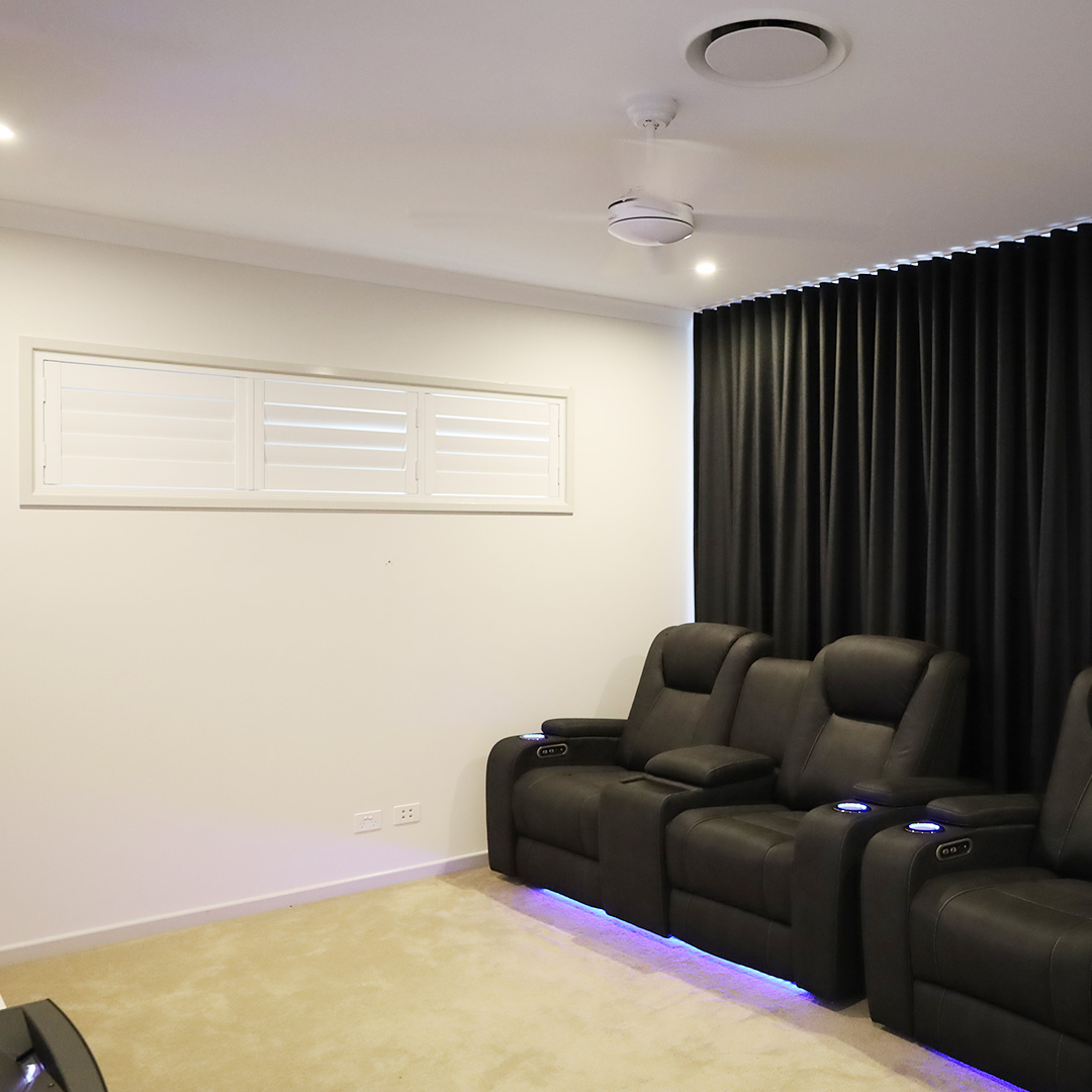 Theatre room featuring a white plantation shutter in a highlight window, next to a wall of black blockout curtains. 