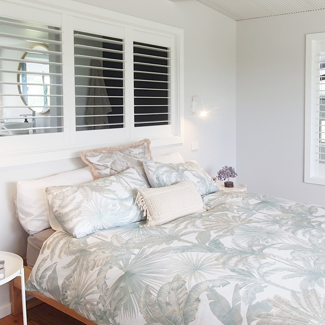 Tropical white loft bedroom featuring a Plantation Shutter above the bed, looking through to the bathroom.