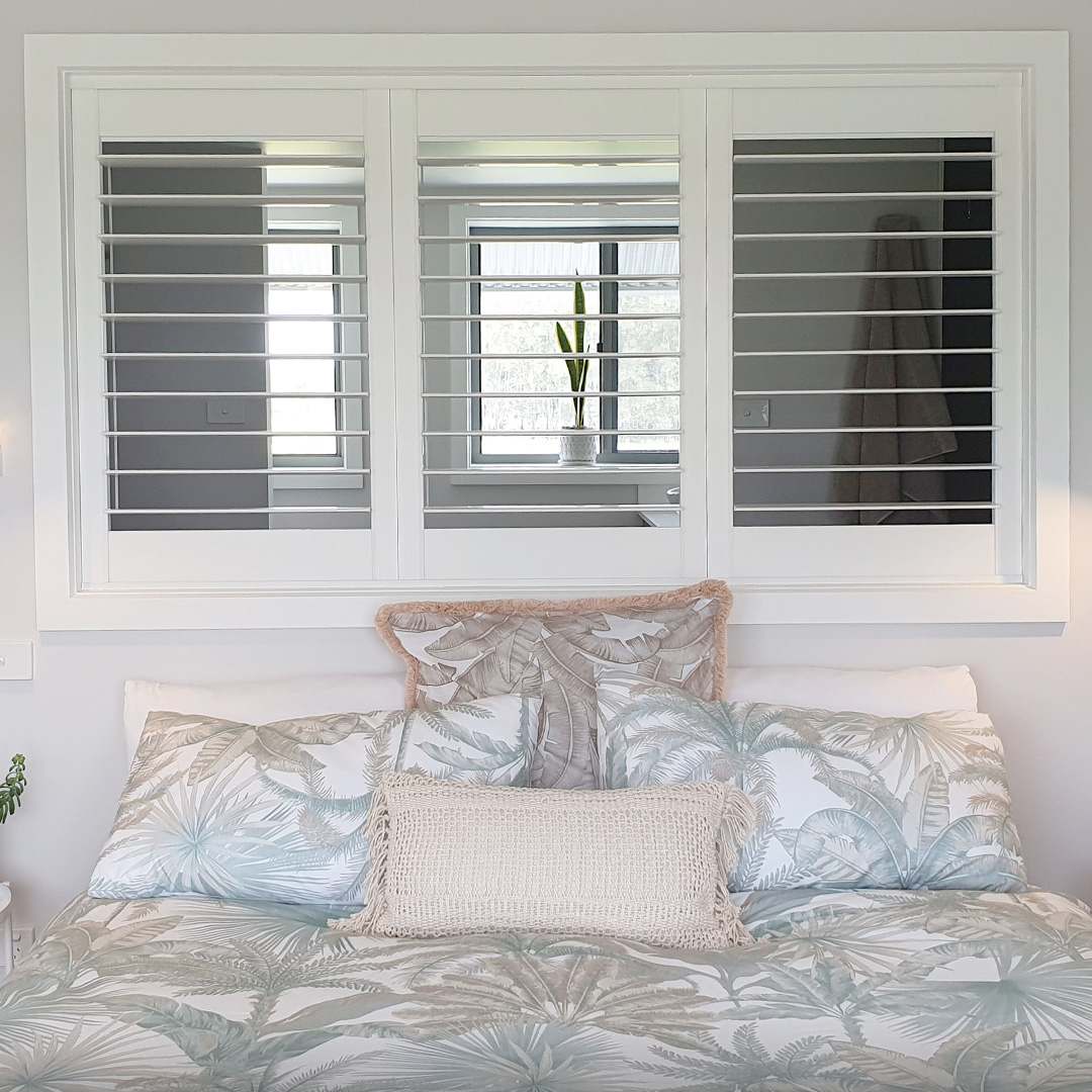 Tropical white loft bedroom featuring a Plantation Shutter above the bed, looking through to the bathroom.