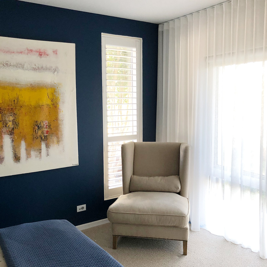 Master bedroom featuring plantation shutters and white sheer curtains with a motorised blockout roller blind behind them. 