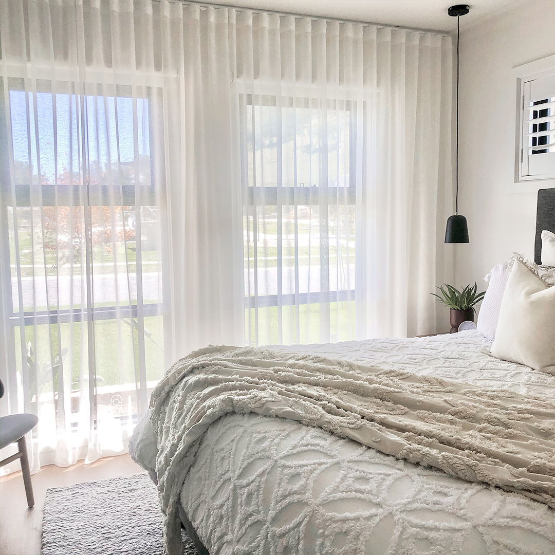 A light and bright bedroom corner showing white sheer curtains on the window, with motorised roller blinds behind. 